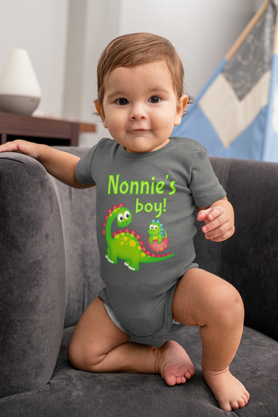 A smiling baby boy wearing a grey onesie with a dinosaur graphic and text that says "Nonnie's boy!"