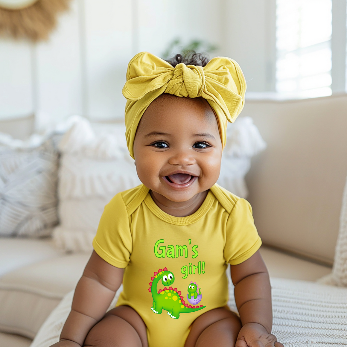 A smiling baby girl wearing a yellow onesie with a dinosaur design and a yellow headband.