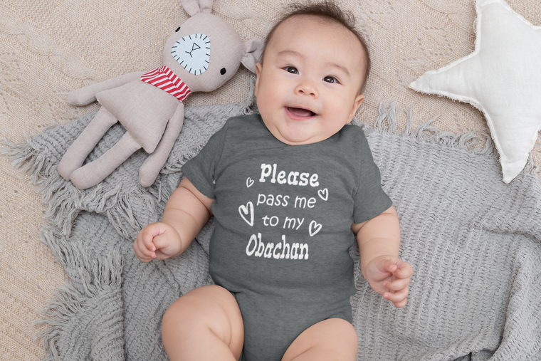 A smiling baby wearing a gray onesie with the text "Please pass me to my Obachan" lies on a gray blanket next to a stuffed animal.