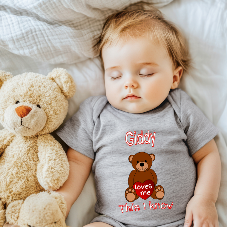 A sleeping baby wearing a gray onesie with a teddy bear and the text "Gippy loves me this I know" printed on it.