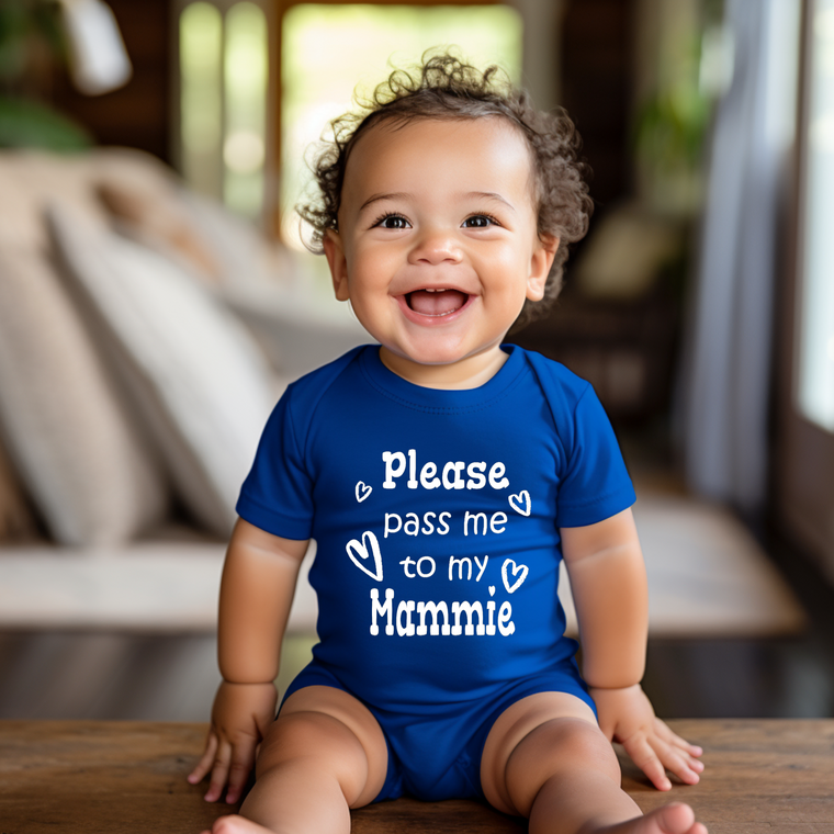 A smiling baby boy wearing a blue onesie with the text "Please pass me to my Mammie" is sitting on a wooden surface.