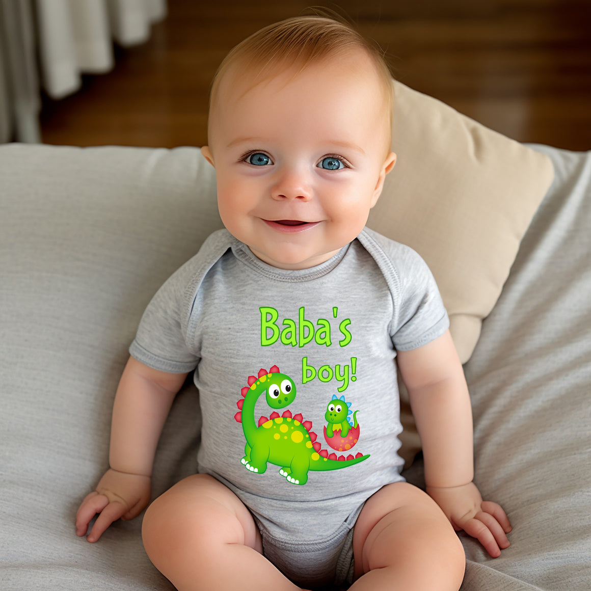 A smiling baby wearing a gray onesie with a dinosaur graphic and the text "Baba's boy!"
