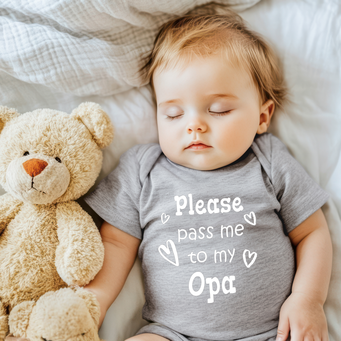A sleeping baby wearing a gray onesie with the text "Please pass me to my Opa" is lying next to a teddy bear on a bed.