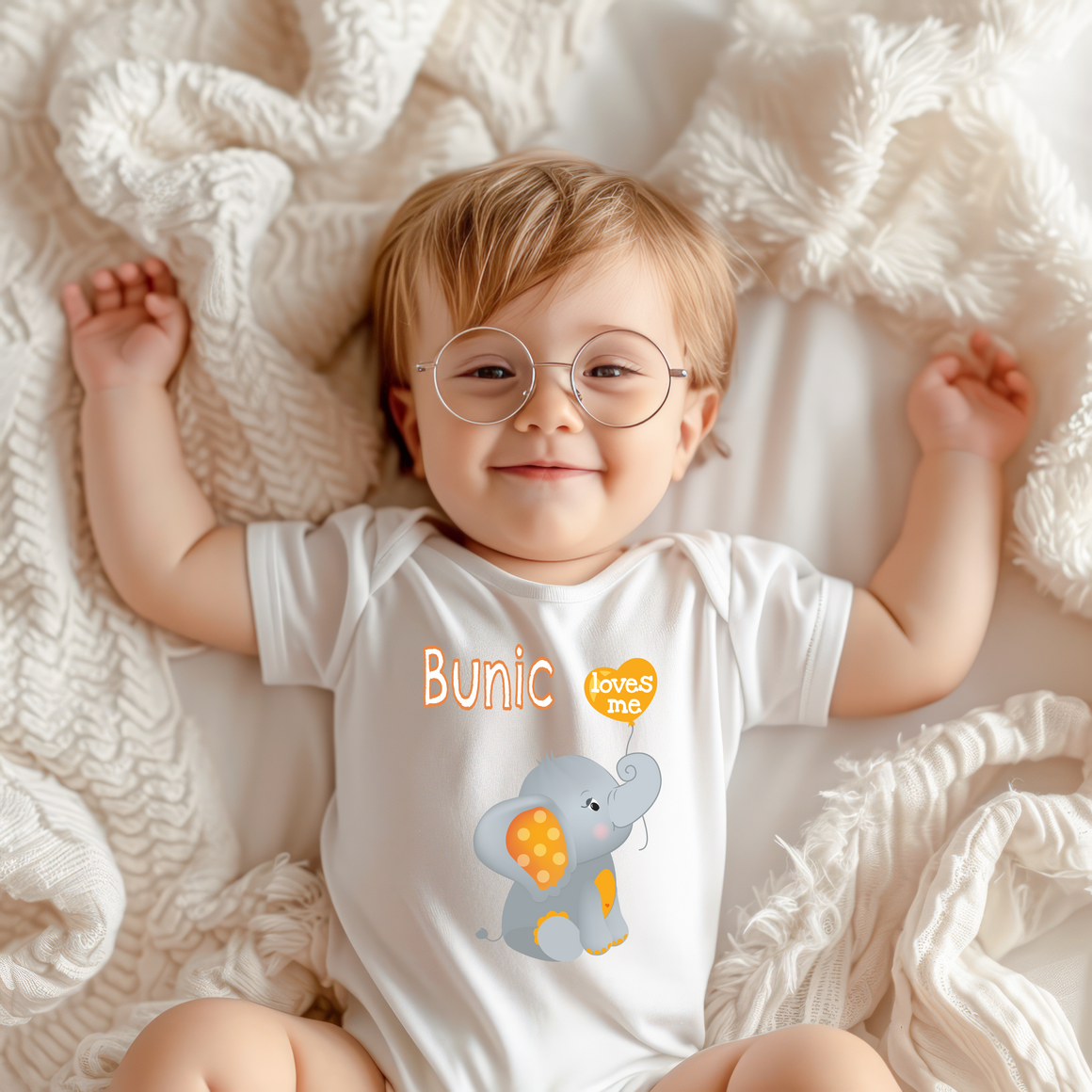 A smiling baby wearing a white onesie with an elephant graphic and the word "Bunic" printed on it, lying on a white blanket.